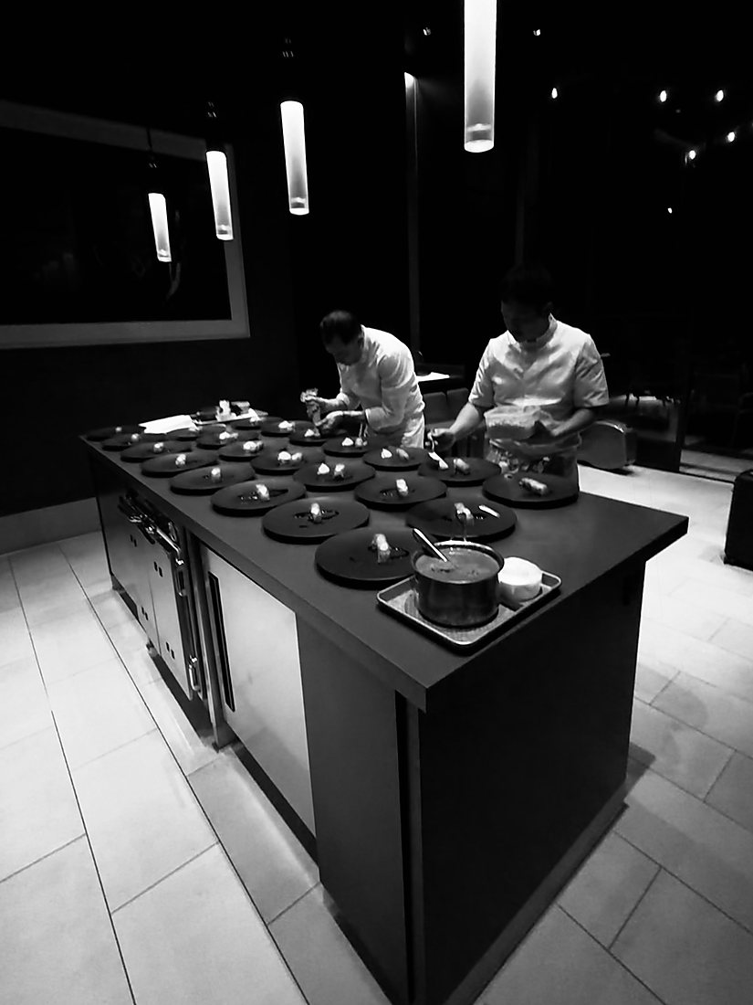 Two cooks in whites plating twenty identical courses along the pass in a darkened kitchen before service